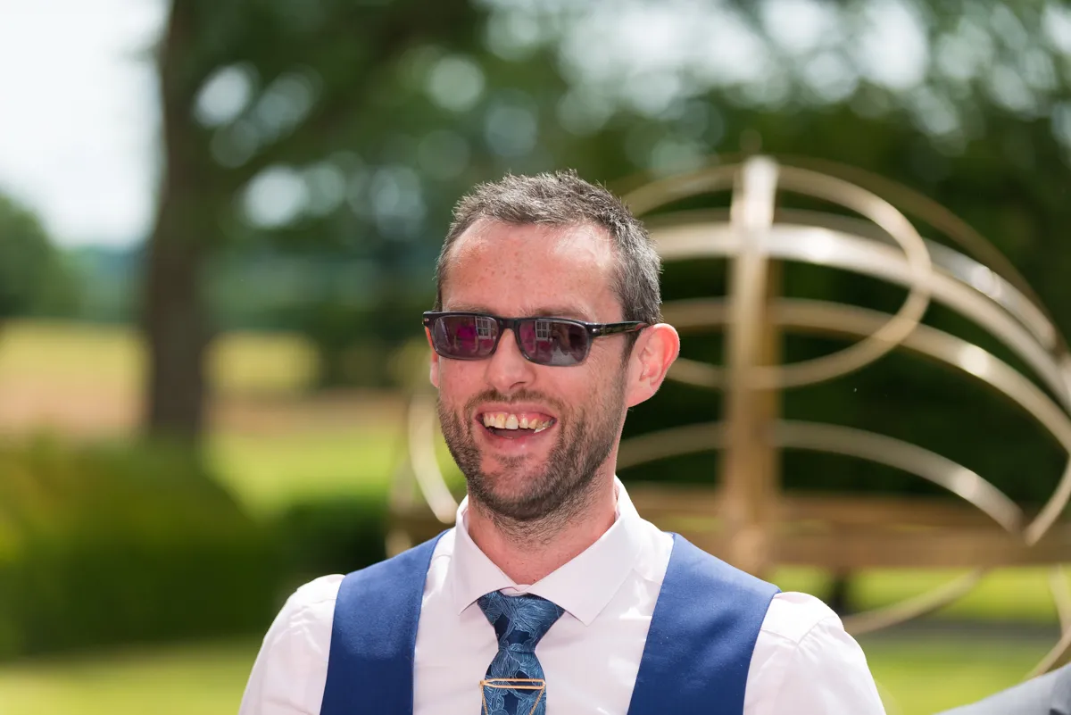 Smiling man wearing sunglasses, a white shirt, blue vest, and patterned tie standing outdoors with blurred greenery and metal sculpture in the background.