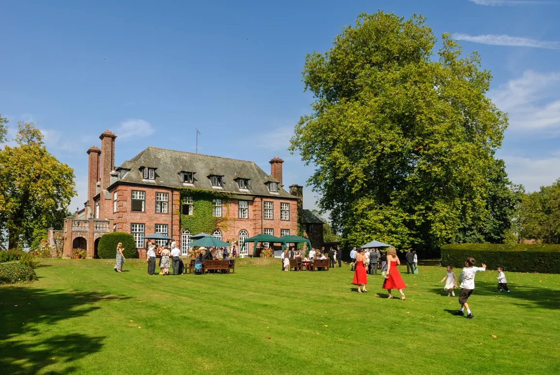 People gathered on the lawn outside a large brick mansion with green ivy and tall trees under a clear blue sky.