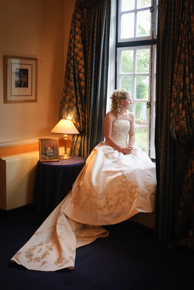 Bride in a strapless white wedding gown sitting on a window ledge looking outside, with dark patterned curtains partially drawn.