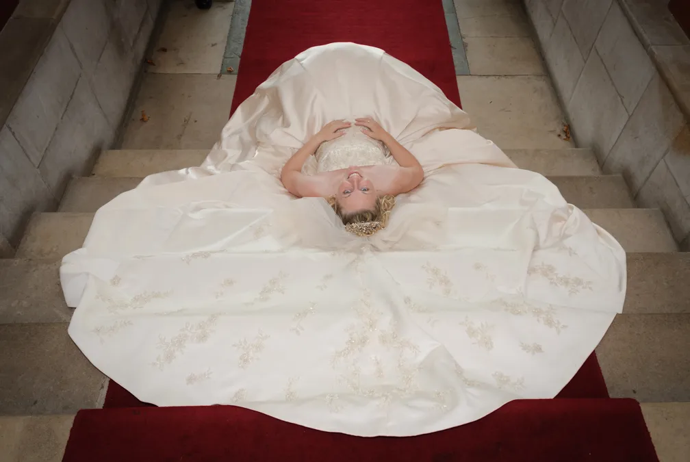 Bride with a floral embroidered white wedding dress lying on stairs with red carpet, smiling upwards.