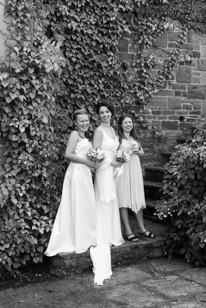 Three smiling females in formal attire stand on stone steps with ivy-covered brick wall background, the two on left in white dresses holding bouquets.