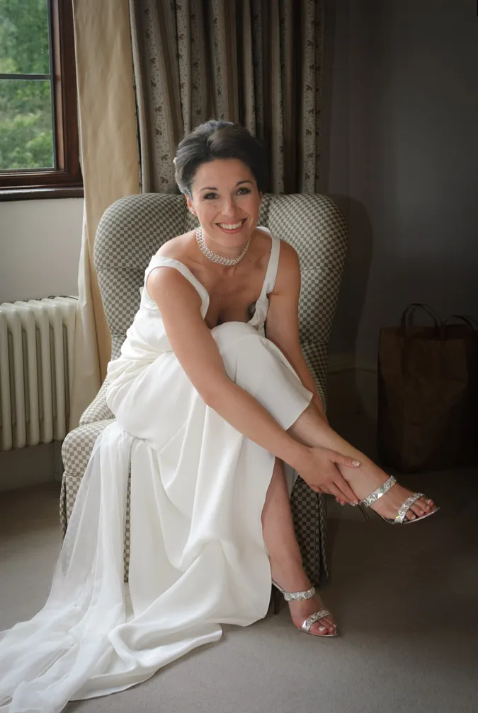 Smiling bride in a white sleeveless gown sitting on a checkered armchair, adjusting her silver high-heeled sandal.