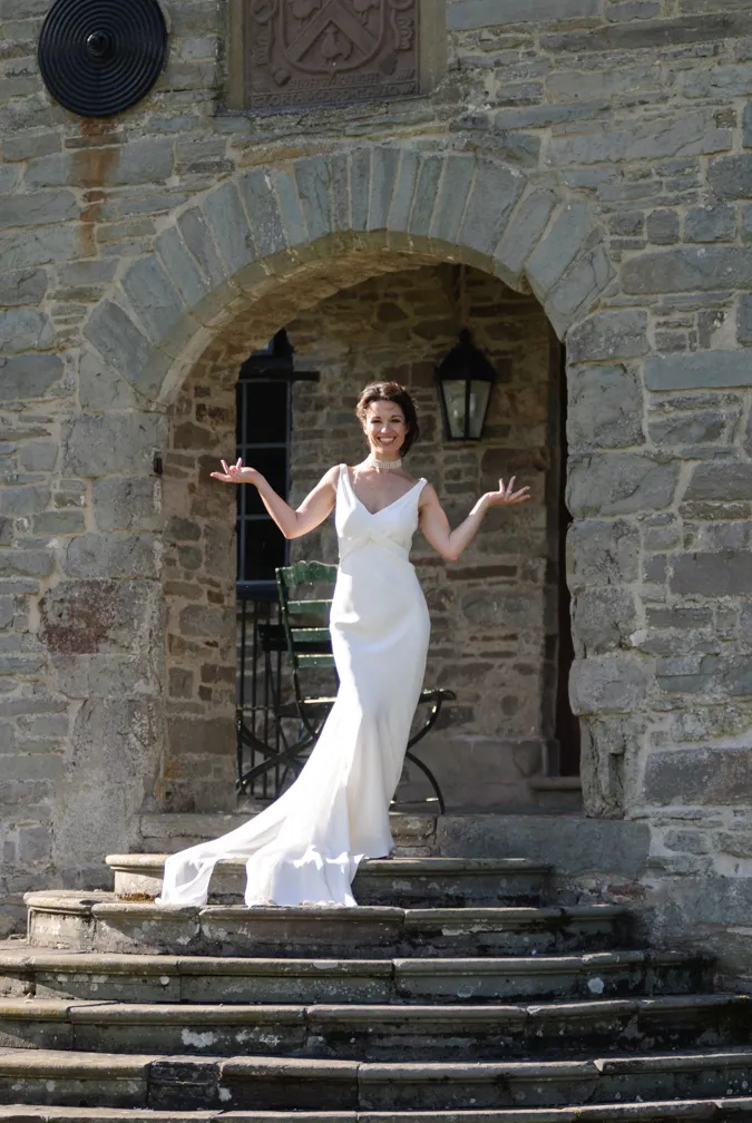 Woman in a white sleeveless gown standing on stone steps under a stone archway, smiling with arms raised.