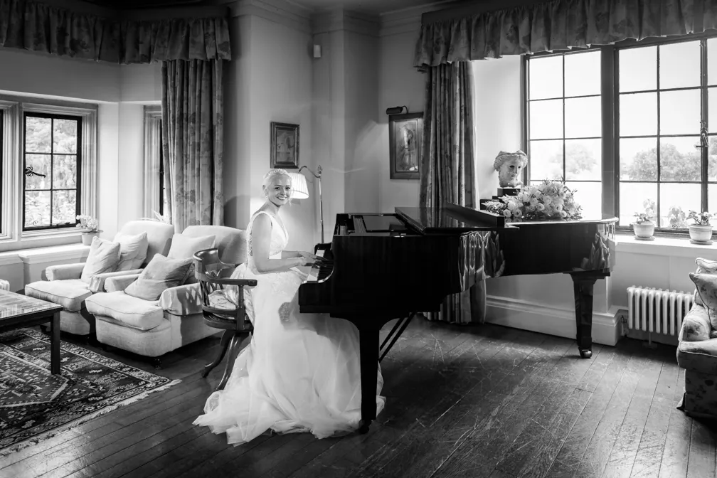 Bride in a wedding dress sitting at a grand piano in a cozy living room with large windows and comfortable chairs.