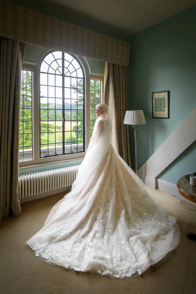 Bride in a lace wedding gown with a long train and veil standing by a window with a scenic outdoor view.