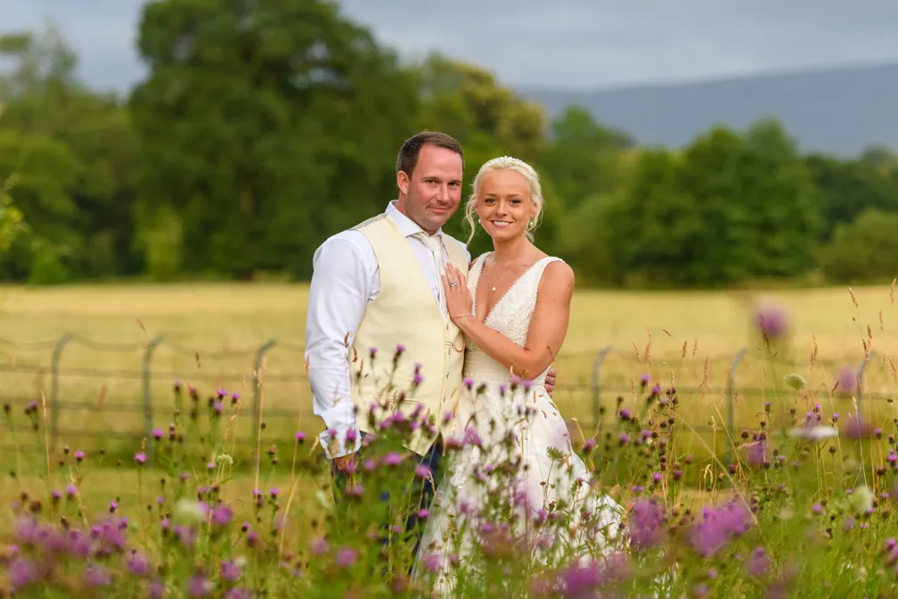 Bride in a white wedding dress and groom in a cream vest standing closely in a field with purple wildflowers and trees in the background.