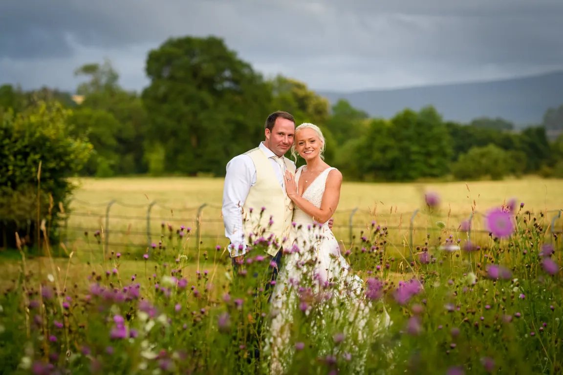 Bride and groom posing together in a field with purple wildflowers and greenery under a cloudy sky.