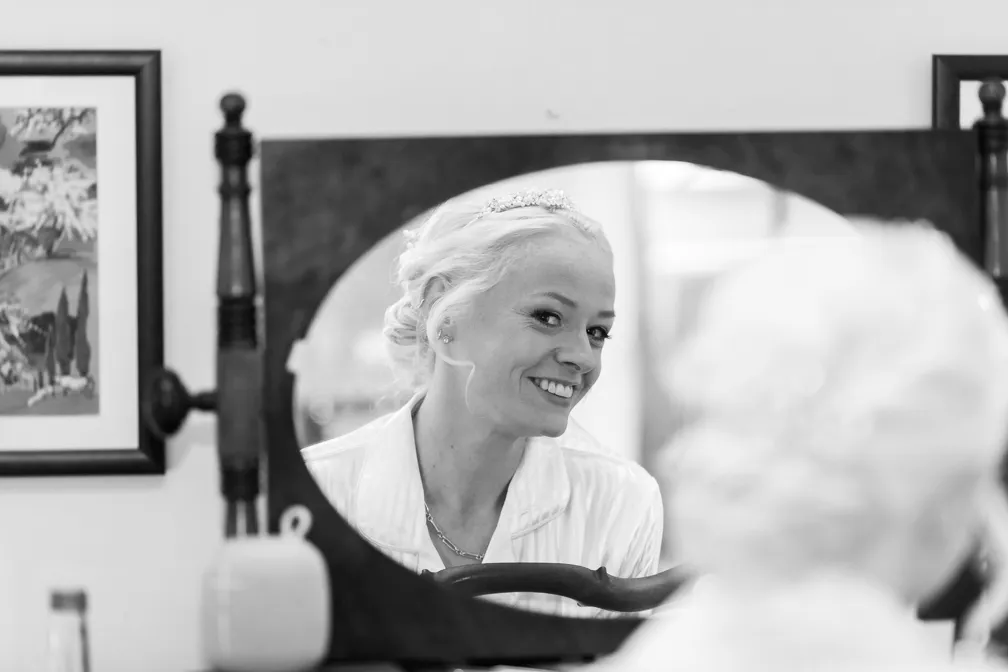 Smiling bride with a tiara and light-colored robe looking at her reflection in a vintage oval mirror.