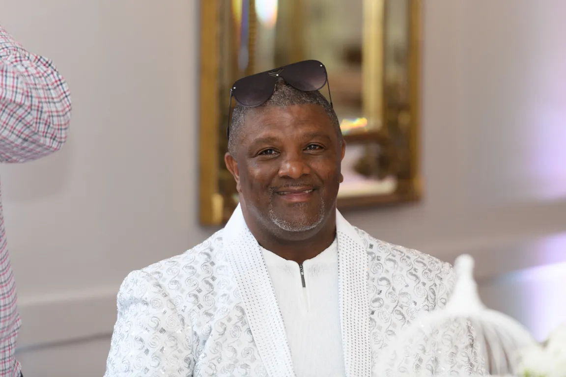 Smiling man wearing a white patterned blazer and sunglasses on his head at an indoor event.