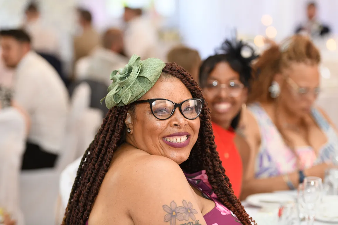 Smiling woman with braided hair, glasses, and green floral headpiece, sitting at a table with others in a bright indoor setting.