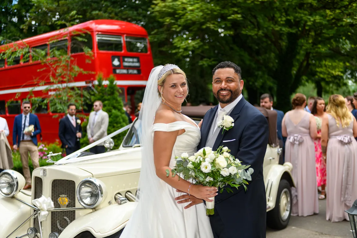 Bride and groom smiling and posing with a bouquet in front of a vintage white car and a red double-decker bus at an outdoor wedding.