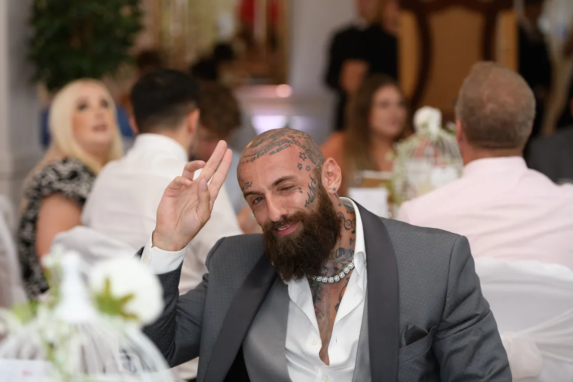 A tattooed man with a beard and diamond necklace in a gray suit winks and makes a peace sign at a formal event.