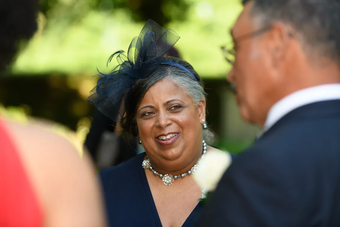 Smiling woman wearing a navy dress, navy fascinator, pearl necklace, and earrings at an outdoor event.
