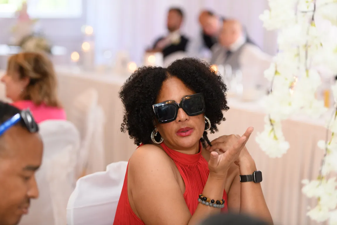Woman with curly hair wearing large black sunglasses and a red sleeveless top gesturing with her hand at an indoor event.