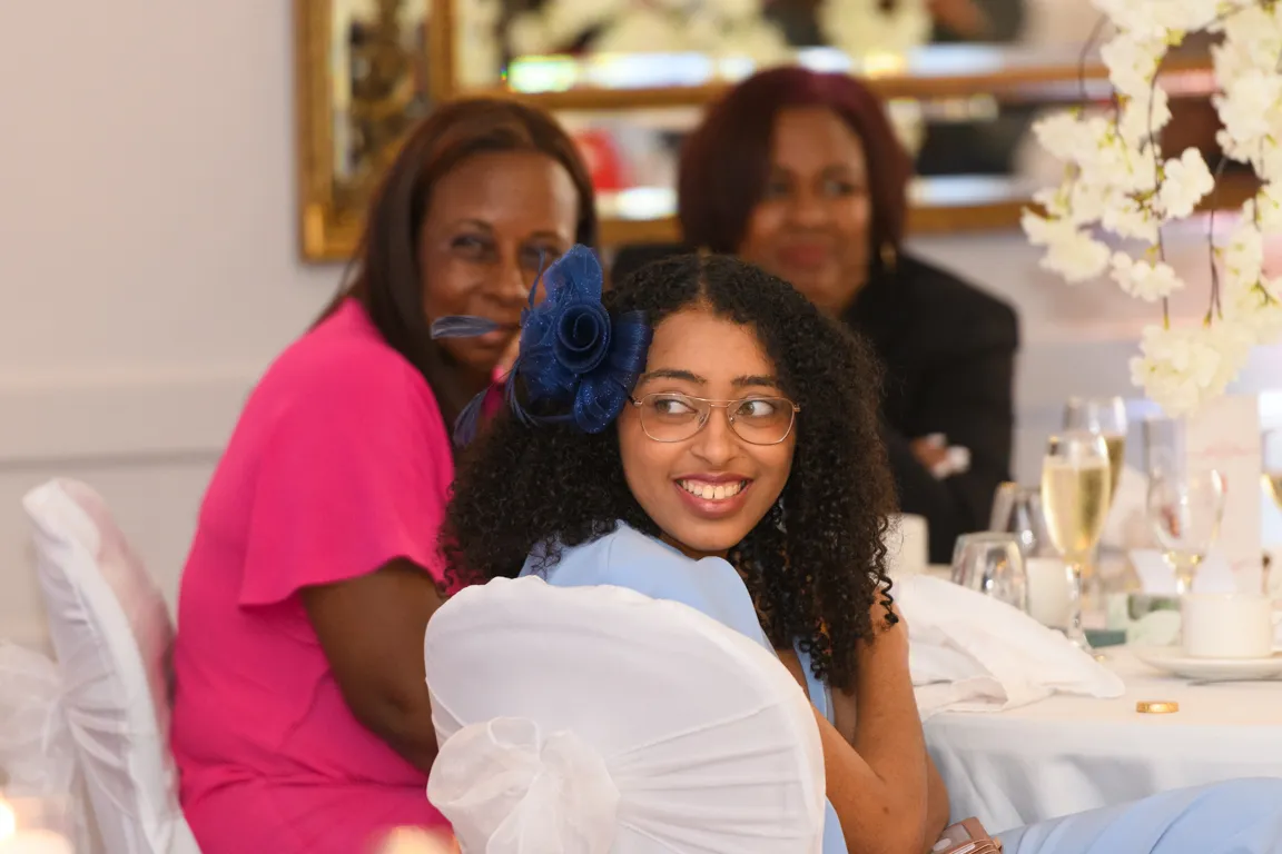 Young woman with curly hair and glasses wearing a blue dress and a large blue flower headpiece, smiling and seated at a decorated table with two women in the background.