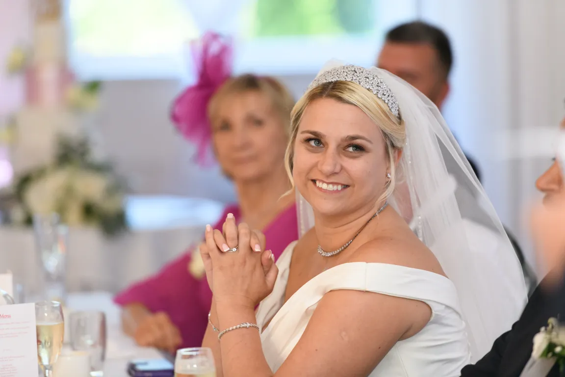 Smiling bride in a white off-shoulder wedding dress with a jeweled headband and veil, seated at a wedding reception table.