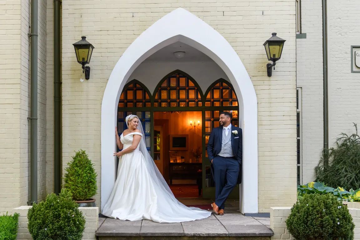 Bride in a white wedding gown and groom in a dark suit standing under a white arched doorway with stained glass windows.