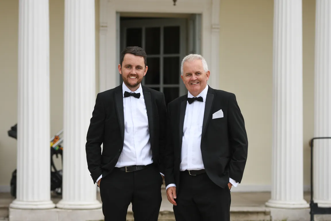 Two men in black tuxedos with bow ties standing side by side in front of white columns and a doorway.