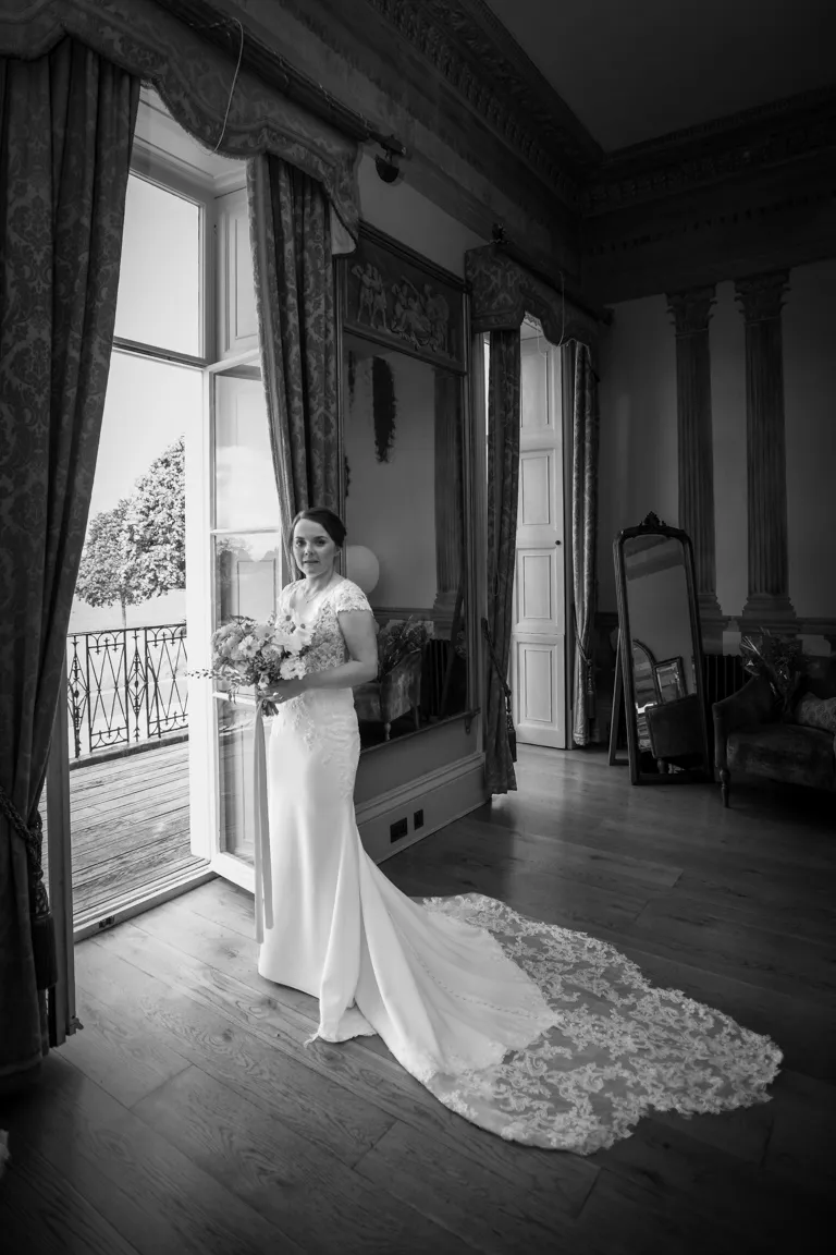 Bride in a lace wedding dress holding a bouquet standing near an open balcony door in an elegant room with tall windows and curtains.