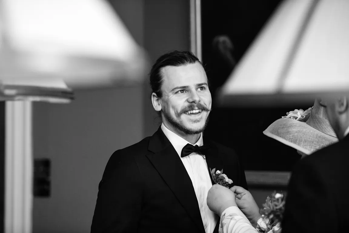 Smiling man with a mustache wearing a tuxedo while someone pins a boutonniere on his lapel in a black and white photo.