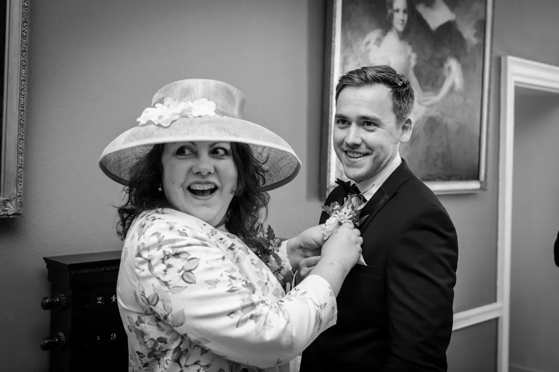 Woman in a floral jacket and large hat smiling while pinning a boutonniere on a smiling man in a suit and bow tie.