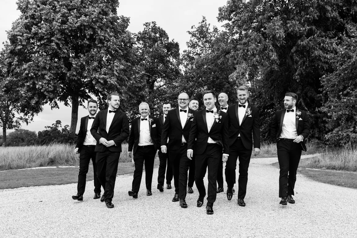 Group of men in tuxedos walking outdoors on a gravel path with trees in the background.
