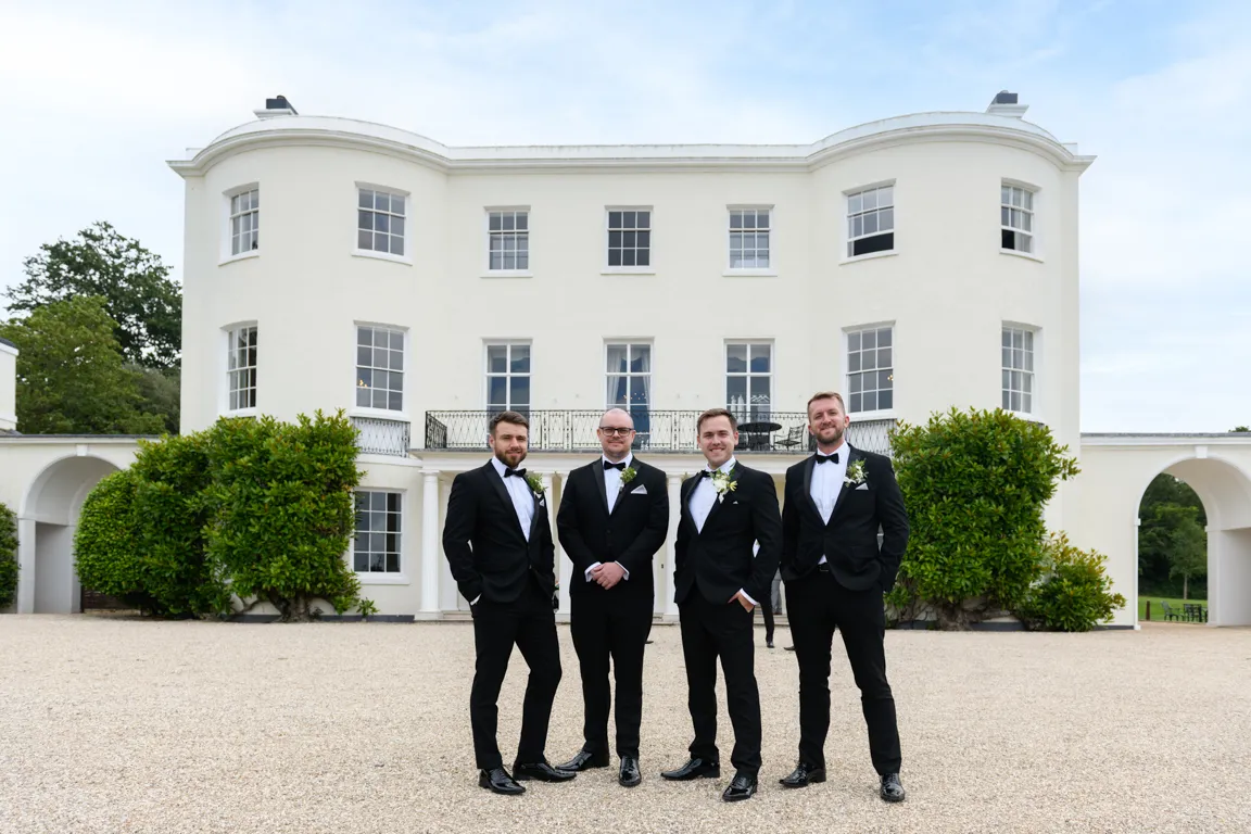 Four men in black tuxedos standing on gravel in front of a large white mansion with greenery on both sides.