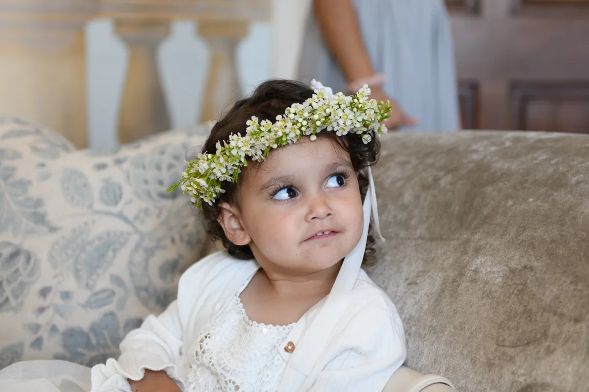 Young child with curly hair wearing a white dress and a flower crown, sitting on a beige couch and looking to the side.