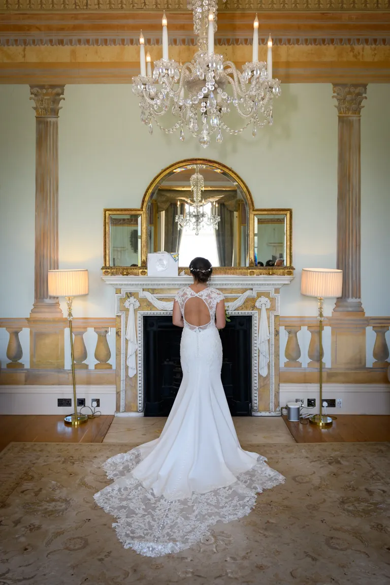 Bride in a white lace wedding dress standing with her back to the camera in an elegant room with a chandelier and ornate fireplace.
