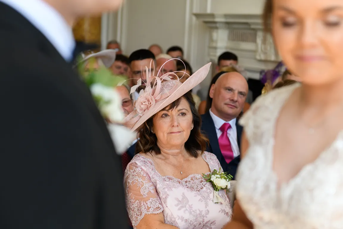Woman in a pink lace dress and matching fascinator with a corsage attentively watching a wedding ceremony indoors.