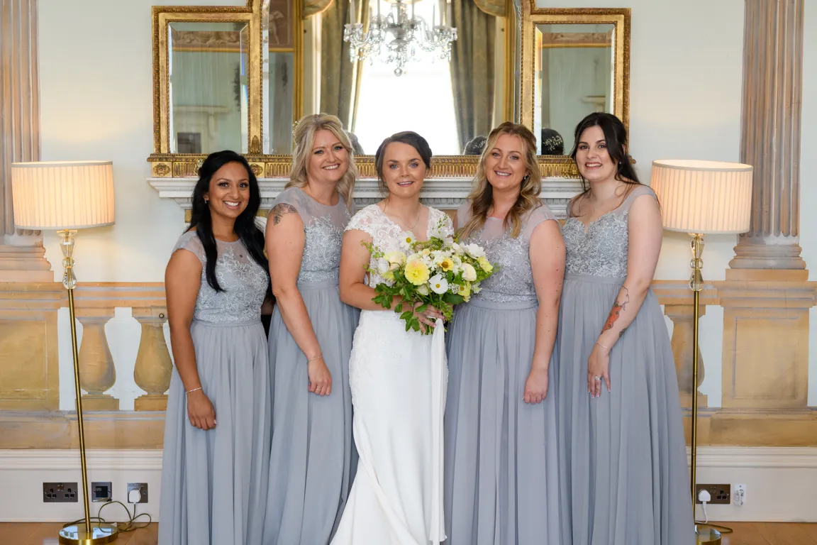 Bride in white wedding dress holding a bouquet of yellow and white flowers, standing with four bridesmaids in matching silver-gray dresses in an elegant room with lamps and mirror.