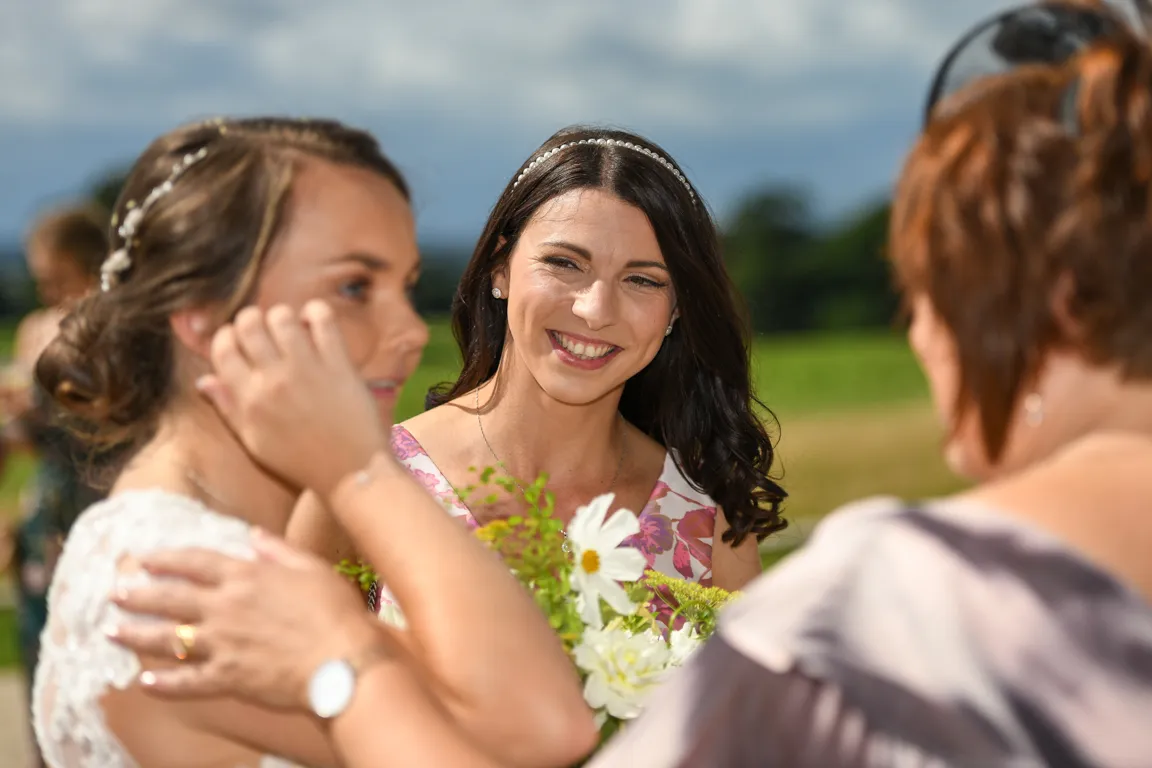Three women interacting outdoors, one holding a bouquet of white flowers, with green fields and cloudy sky in the background.