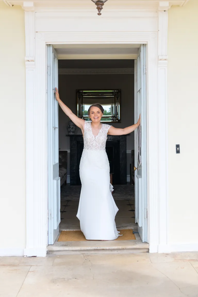 Smiling bride in a white lace wedding dress standing in an open doorway with arms raised holding the door frames.