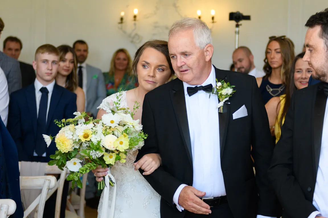 Bride in white lace dress holding yellow and white flower bouquet walks arm-in-arm with an older man in a black tuxedo during wedding ceremony.
