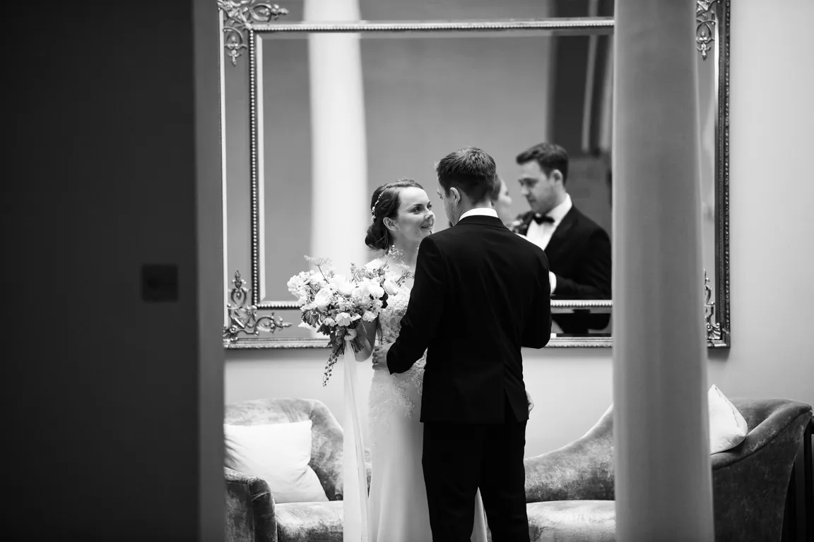 Bride and groom standing close, facing each other, with bride holding a bouquet in front of a large ornate mirror reflecting a man in a tuxedo.