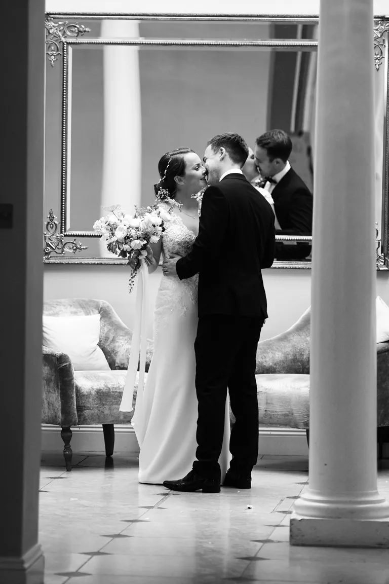 Black and white photo of a bride and groom sharing a close, intimate moment in front of a large decorative mirror, with the bride holding a bouquet.