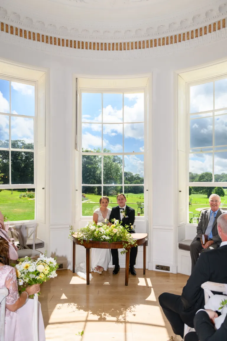 Bride and groom sitting together at a table decorated with flowers in a bright room with large windows overlooking a green garden.