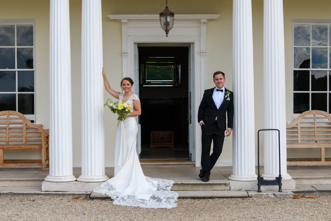 Bride in white lace wedding gown holding bouquet standing by pillar and groom in black tuxedo leaning on adjacent pillar outside a building entrance.