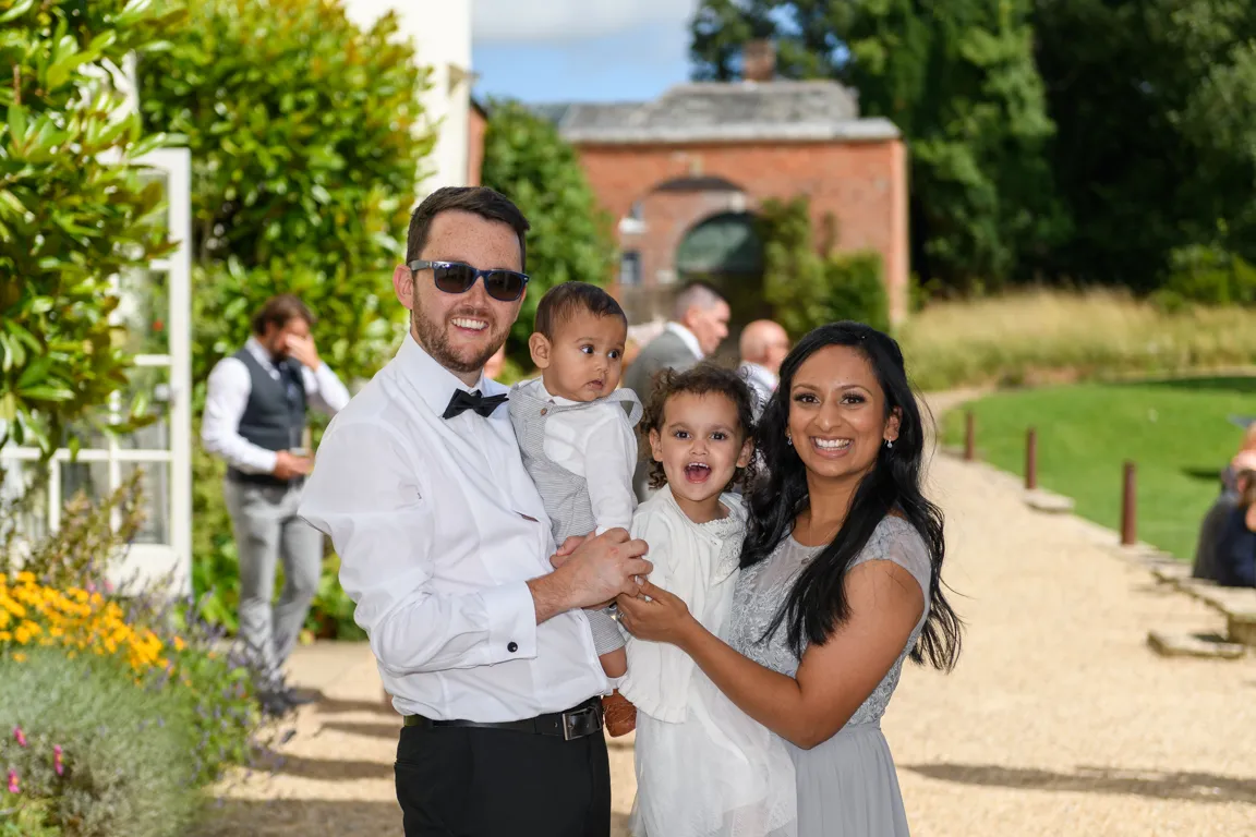 Smiling family of four posing outdoors with a man in a white shirt and bow tie holding a baby and a woman in a gray dress holding a toddler.