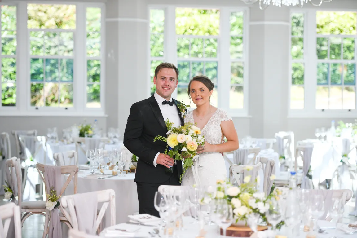 Bride in white lace dress and groom in black tuxedo standing indoors with wedding tables and floral decorations in the background.