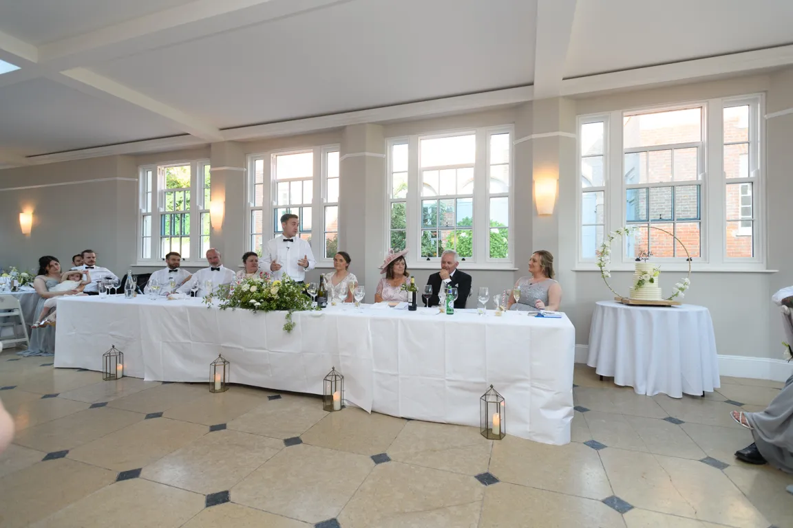 Wedding party seated at a long table with white cloth, with a man standing and giving a speech in a bright room with large windows.