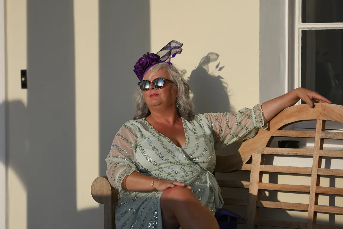 A woman wearing sunglasses, a purple floral headpiece, and a light green sequin dress sits on a wooden bench against a light wall with shadows.