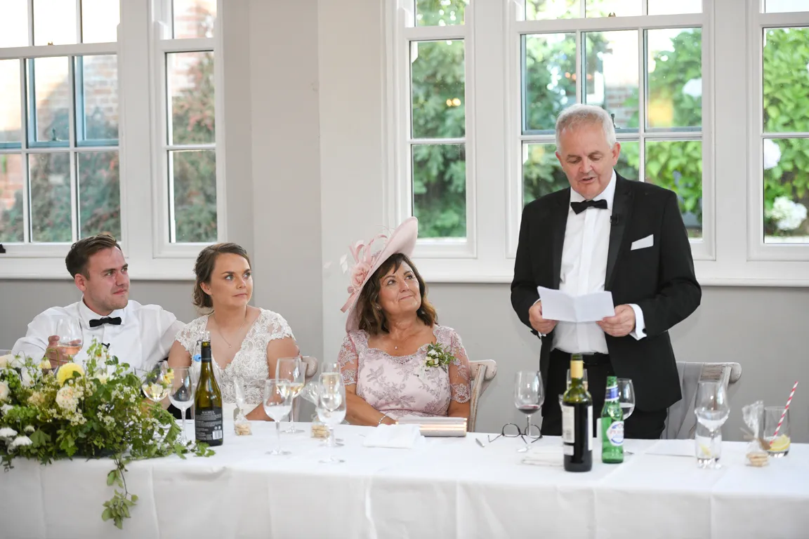 A man in a tuxedo giving a speech at a wedding, while a bride, groom, and woman in a pink dress with a matching hat listen at a table with wine glasses and floral arrangements.