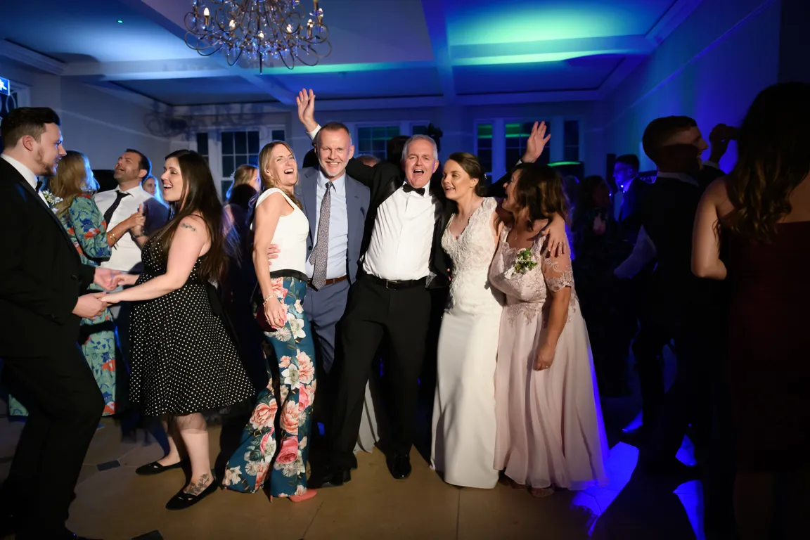 Group of people smiling and dancing indoors at a wedding reception, with the bride in a white dress and guests surrounding her.