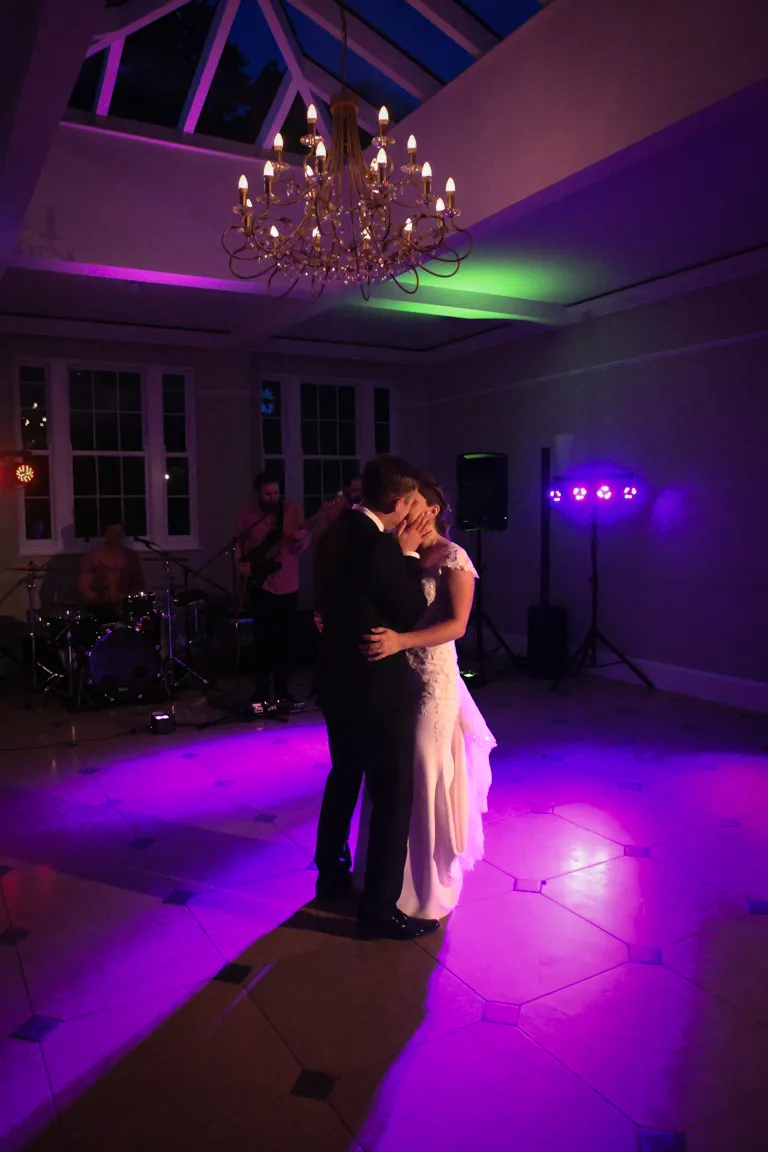 Bride and groom sharing a kiss while dancing under a chandelier with purple and green lighting in a reception room.