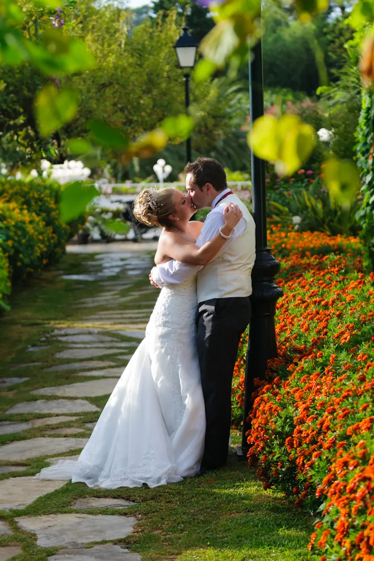 Bride and groom embracing and kissing beside a lamppost with vibrant orange flowers and greenery in a garden.