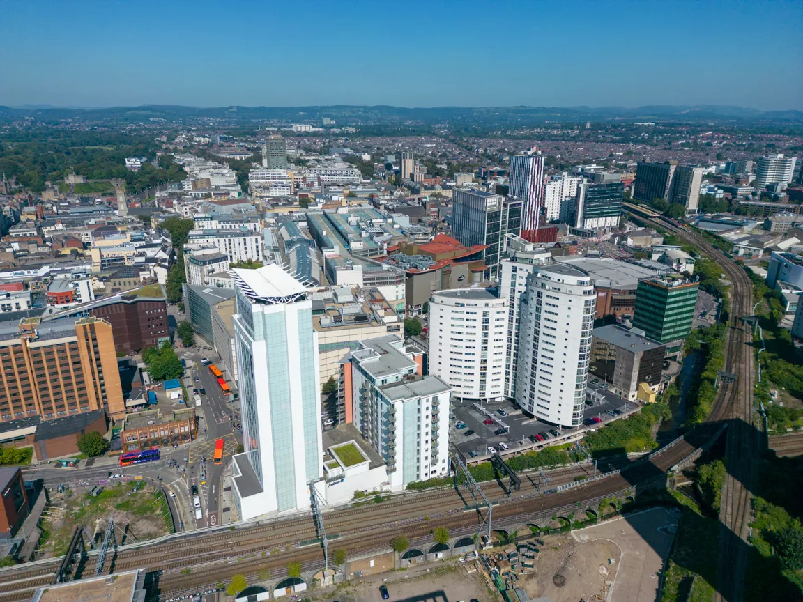 Aerial view of a cityscape with modern high-rise buildings, train tracks in the foreground, and surrounding urban area under a clear blue sky.