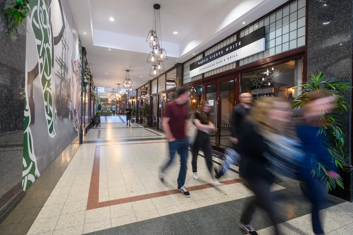 Blurred people walking inside a shopping arcade with a visible sign for Marco Pierre White Steakhouse Bar & Grill.