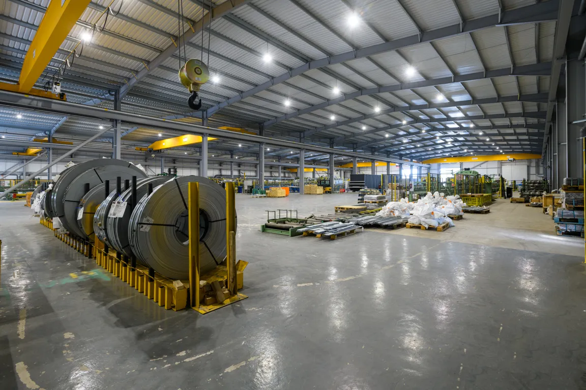 Spacious industrial warehouse with metal coils and pallets of materials under bright ceiling lights and yellow overhead cranes.