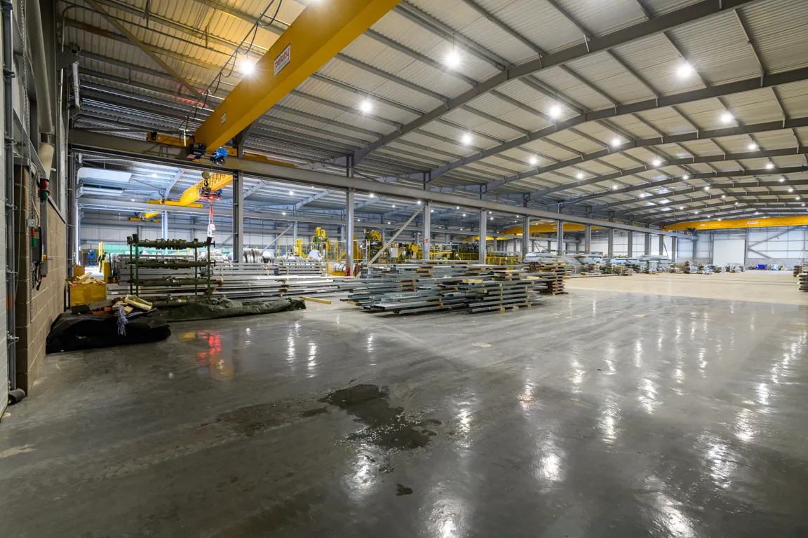 Large industrial warehouse interior with stacks of metal sheets and yellow overhead cranes under bright ceiling lights.